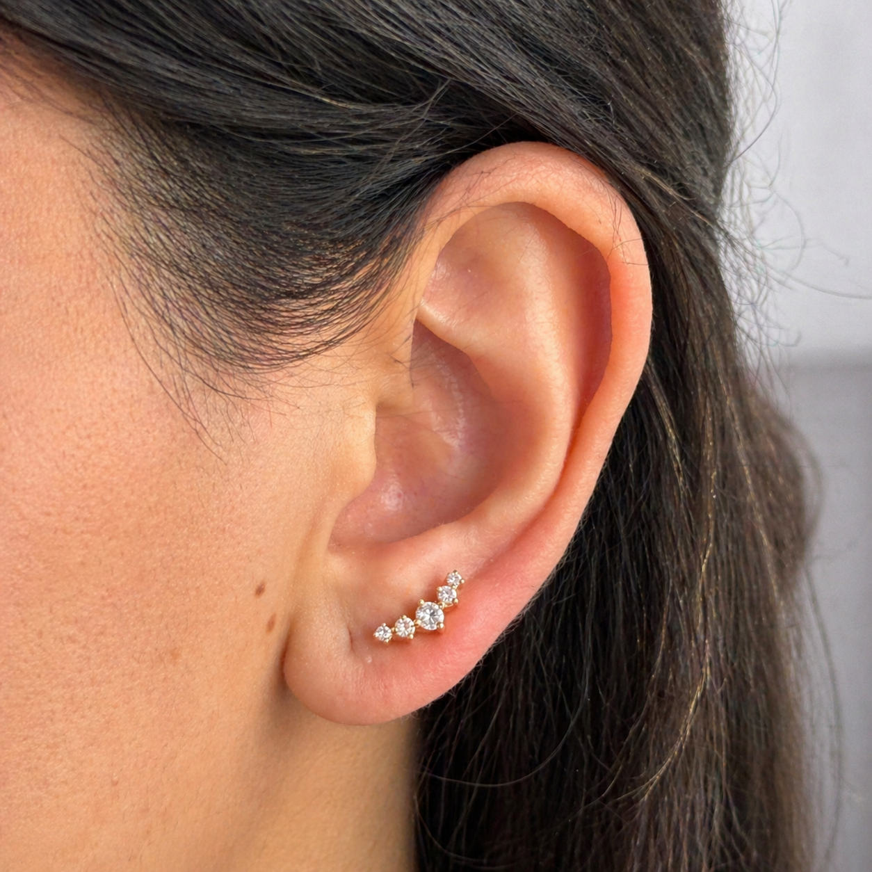 Close-up of an ear wearing a diamond earring with dark hair and neutral background